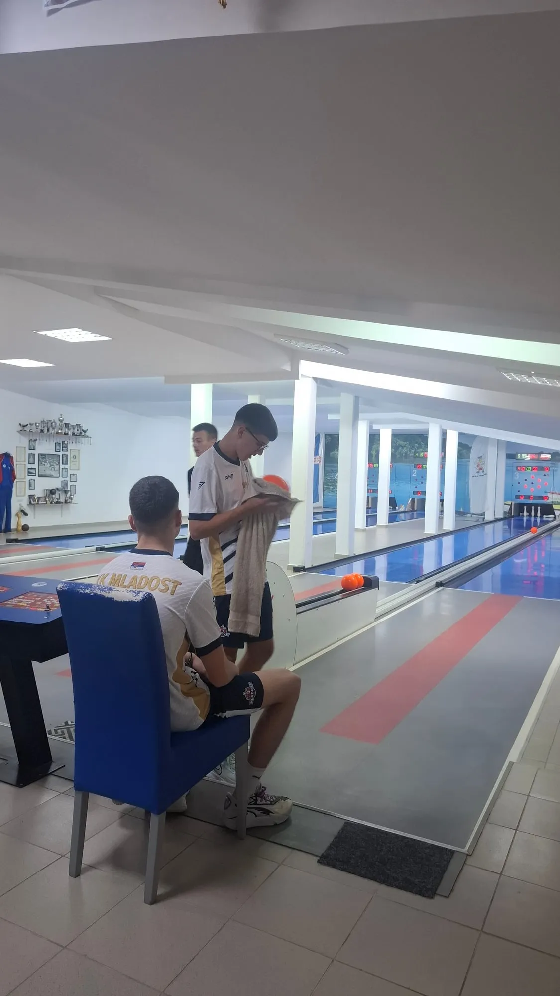 Strahinja Denčić u belom dresu Mladosti briše kuglu peškirom, ispred njega sedi drugi igrač Mladosti.