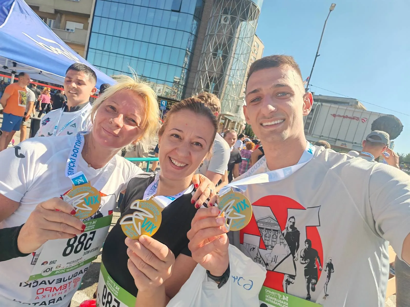 Troje trkača se smeje u kameru i pokazuje medalje 14. Kragujevačkog polumaratona, iza je staklena zgrada.