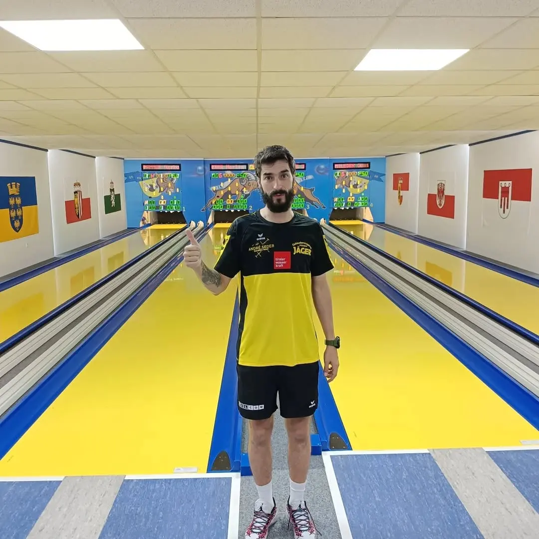 Miloš Simijonović u žuto-crnom dresu podiže palac na kuglani sa žutim stazama, na zidovima su zastave austrijskih pokrajina.