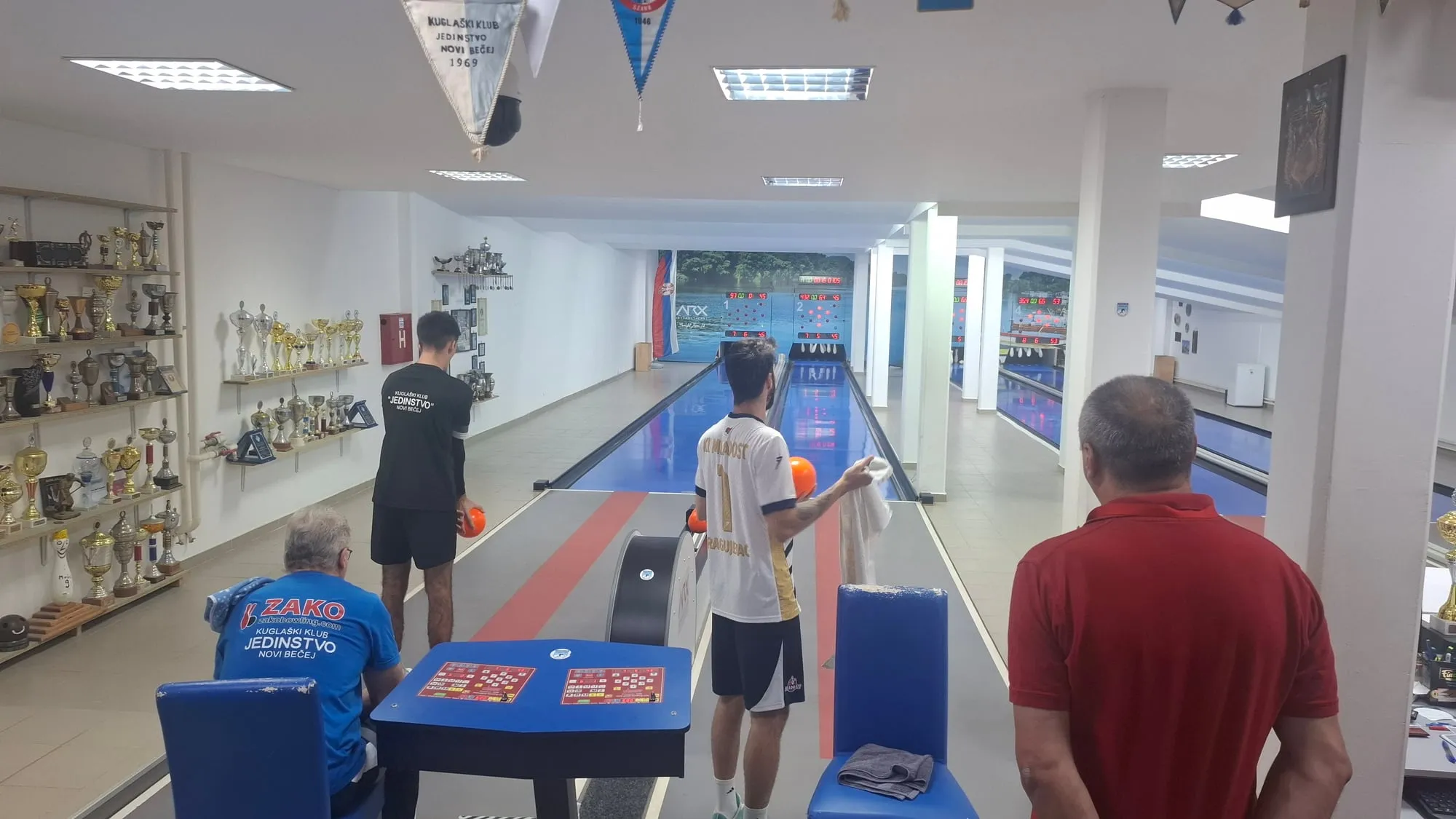 Igrač Jedinstva u crnoj majici i igrač Mladosti u beloj majici sa brojem 1 sa kuglama na stazama 1 i 2, u prvom planu sto za upis i gledaoci.