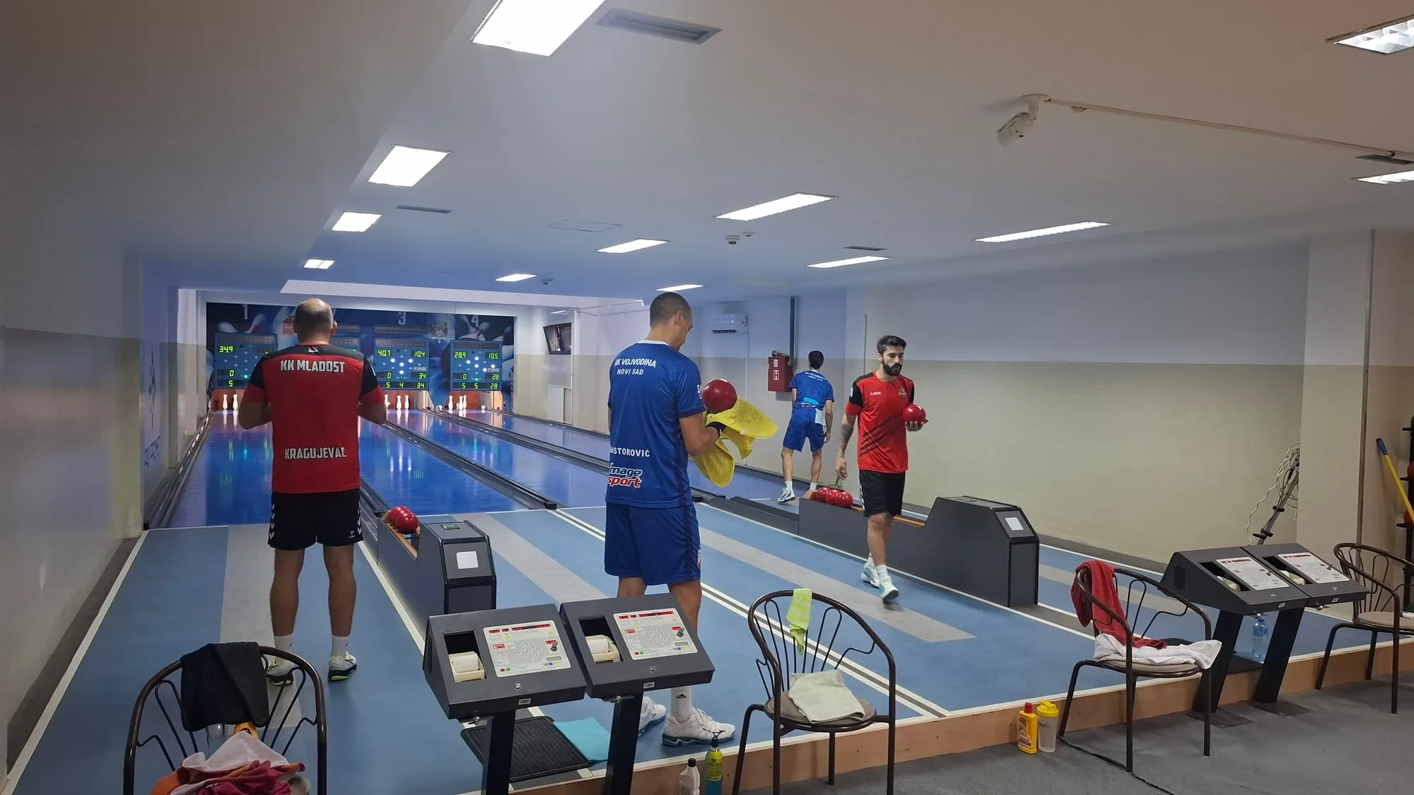 Četiri igrača na stazama: levo igrač u crveno-crnoj majici okrenut stazi, u sredini igrač u plavoj majici briše kuglu, desno igrač u crveno-crnoj majici nosi kuglu, u dnu igrač u plavom.