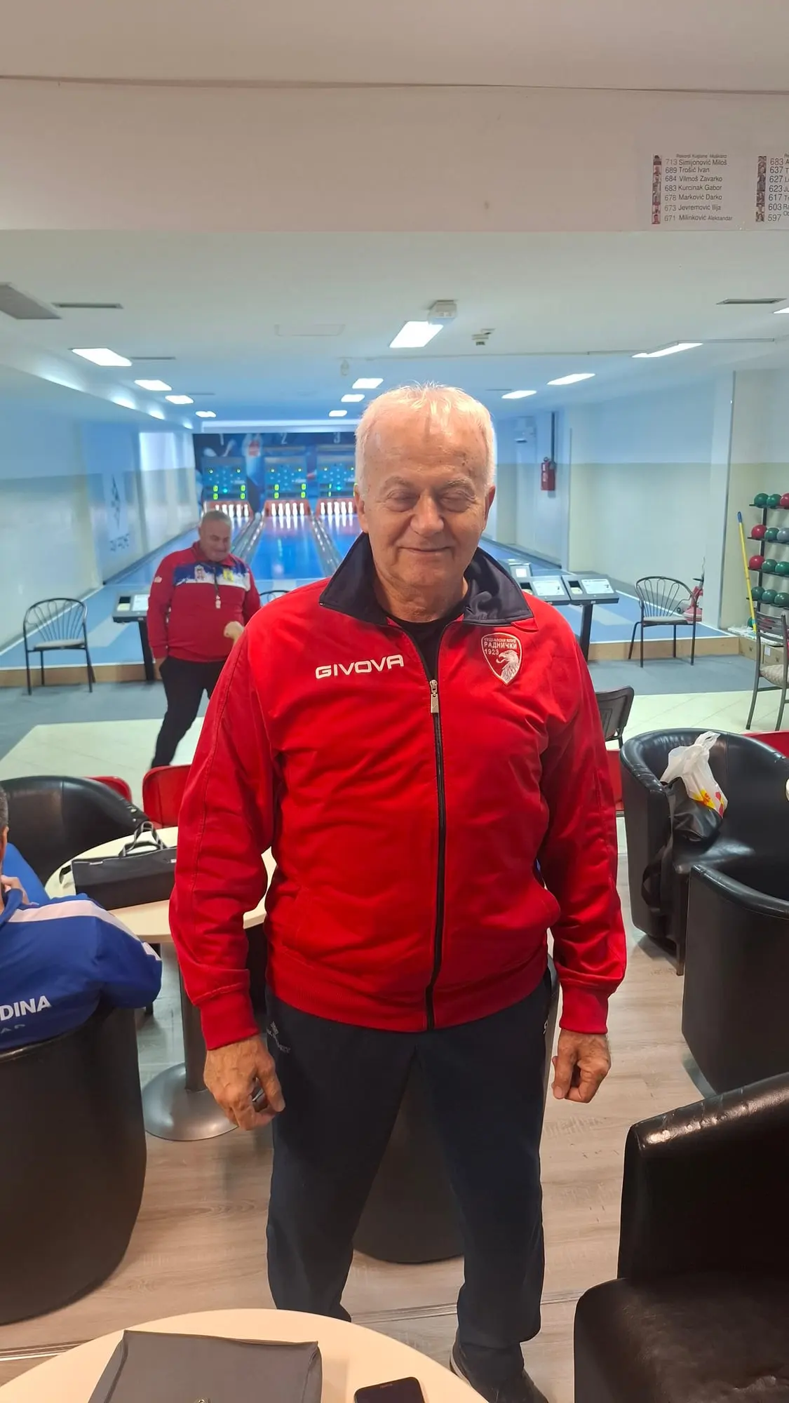 Stariji muškarac u crvenoj jakni stoji u salonu kuglane, iza su staze i semafori, a gore desno tabla sa rekordima.