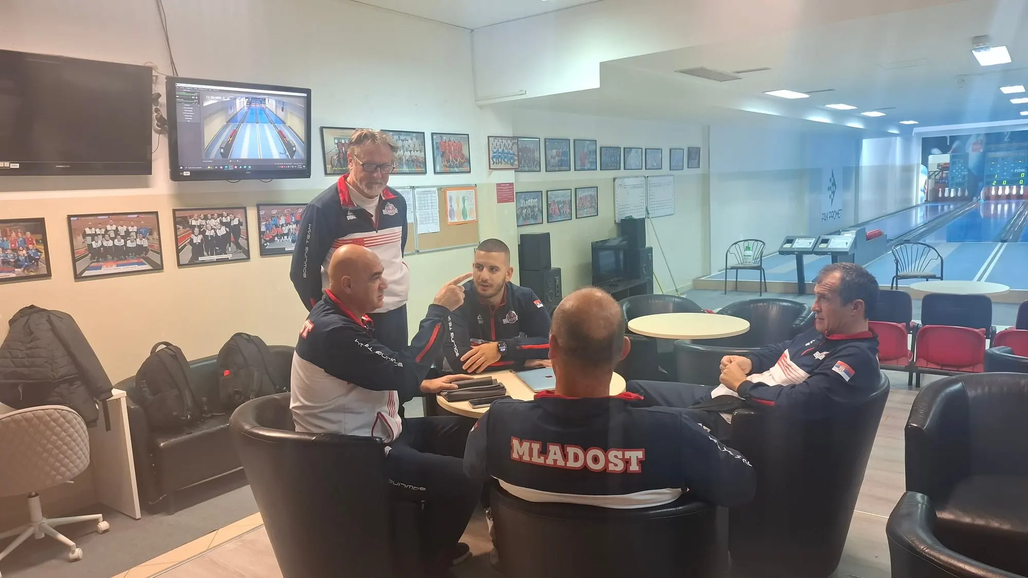 Igrači u teget-belim trenerkama Mladosti sede u foteljama oko stola i razgovaraju, jedan stoji iza njih.