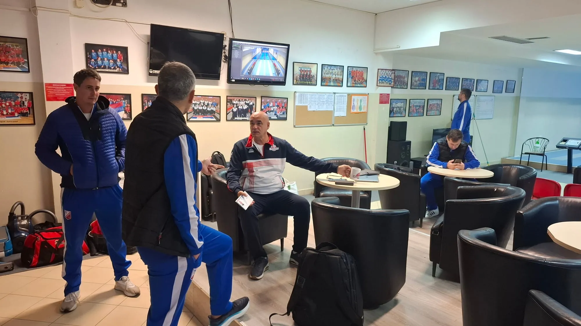 Dvojica u plavim trenerkama Vojvodine stoje i razgovaraju sa čovekom u trenerci Mladosti koji sedi sa šoljom u ruci.