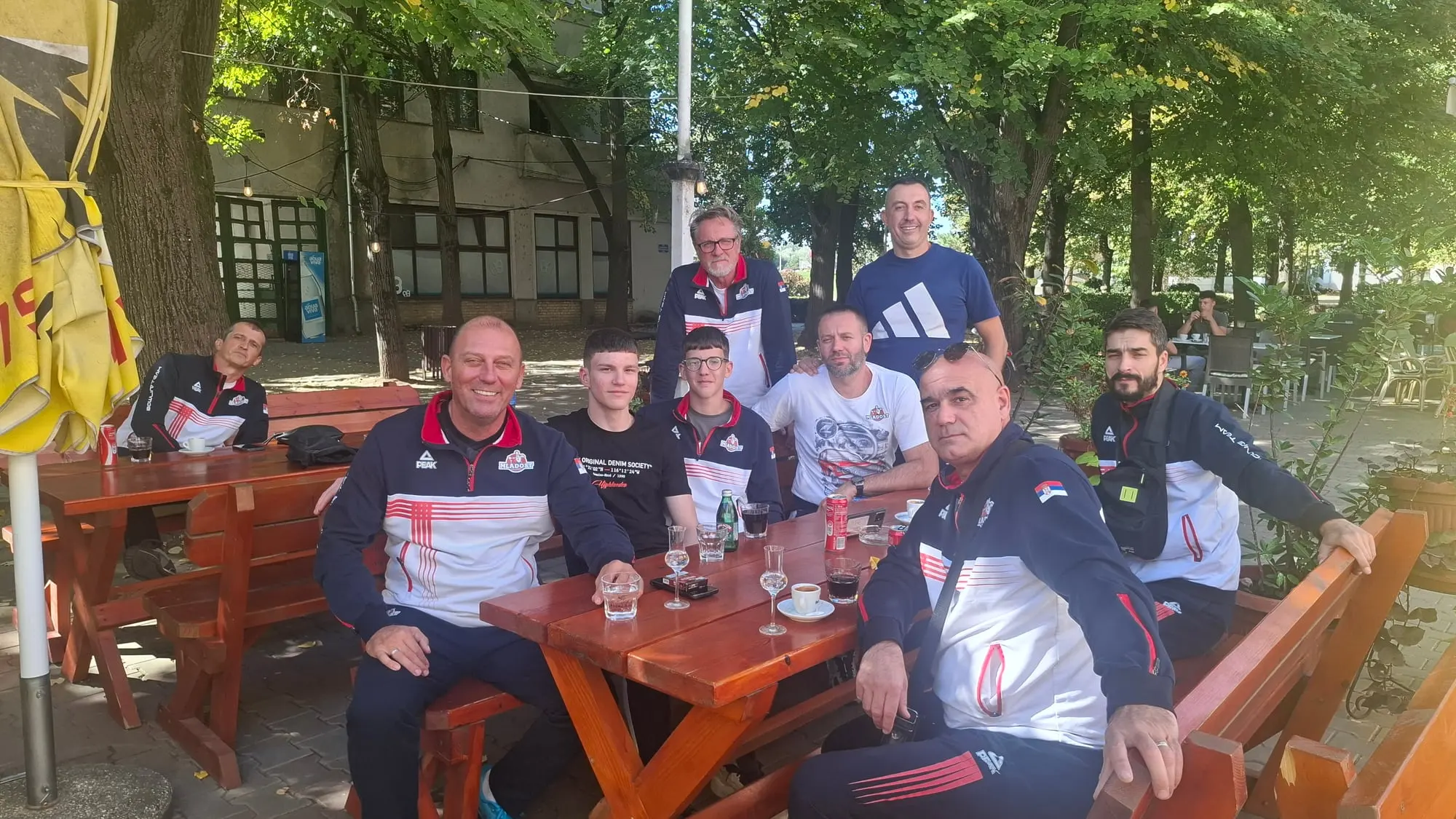 Igrači Mladosti u trenerkama kluba sede za drvenim stolom u bašti kafića, u hladu drveća.